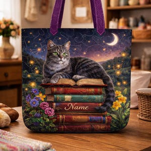 May include: A tote bag featuring a gray tabby cat with green eyes resting on a stack of books. The bag has a night sky design with a crescent moon and stars, and purple handles. The word "Name" is printed on the books.