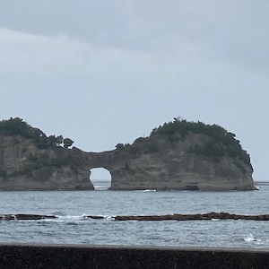 Pode incluir: Uma formação rochosa insular que emerge do oceano, apresentando um arco natural distinto esculpido no seu centro. A massa terrestre é coberta por manchas de vegetação verde e rodeada por águas escuras e agitadas sob um céu nublado.