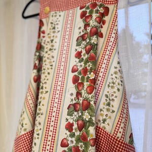 May include: An apron featuring a vertical pattern of red strawberries, green leaves, and white flowers on a cream background, accented by red and white gingham check panels and light blue stripes. The garment has yellow buttons at the shoulder straps.