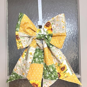 May include: A large, decorative bow with a sunflower and polka dot pattern. The bow is made of fabric and features yellow, green, and cream colors. It is designed to hang on a door or wall, adding a touch of rustic charm.