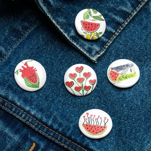 Könnte beinhalten: Fünf runde, weiße Emaille-Anstecknadeln mit farbenfrohen Illustrationen. Die Designs zeigen Wassermelonen, Herzen und Gebäude. Die Anstecknadeln sind auf einer blauen Jeansjacke angebracht.