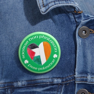 May include: A green button with the words "Free Palestine" and "Saoirse don phalaistin" in white text. The button features a heart-shaped design with the Palestinian flag and the Irish flag, along with shamrock accents.