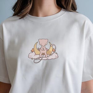 Könnte beinhalten: Cremefarbenes T-Shirt mit einem gestickten Design. Das Design zeigt eine hellrosa Kreatur, die auf einem Halbmond und einer Wolke sitzt. Das Design ist hellbraun umrandet.