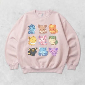 Op de afbeelding: Een lichtroze sweatshirt met negen kleurrijke cartoonfiguren. De karakters zijn gerangschikt in drie rijen van drie. Elk karakter heeft een uniek ontwerp en kleurenschema, waaronder blauw, bruin, geel, roze, groen, zwart en paars.