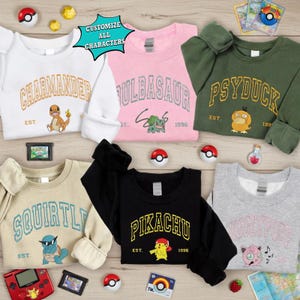 May include: A collection of sweatshirts in various colors, each featuring a different Pokémon character's name in a collegiate font. The sweatshirts include Charmander, Bulbasaur, Psyduck, Squirtle, Pikachu, and Wigglytuff. Additional Pokémon-themed accessories are scattered around.