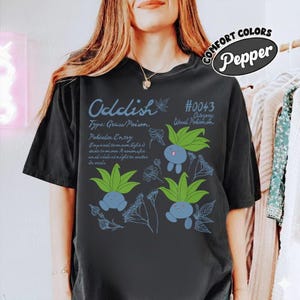 Puede incluir: Camiseta negra con un gráfico de Oddish azul y verde y texto. El diseño incluye el texto "Oddish", "Type: Grass/Poison" y "#0043 Category: Weed Pokemon". La camiseta también tiene las palabras "COMFORT COLORS Pepper".