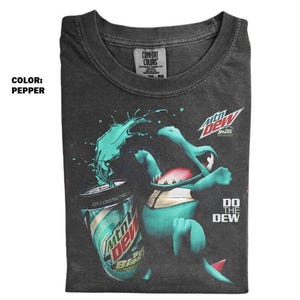 May include: Pepper-colored t-shirt featuring a cartoon creature with a green and teal design, drinking from a Mtn Dew Baja Blast can. The shirt has the text "DO THE DEW" on the lower right.