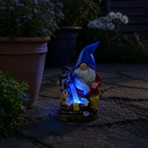 Puede incluir: Una estatua de gnomo de jardín con un gorro azul, una camisa roja y pantalones amarillos. El gnomo sostiene una regadera con luces LED azules que parecen agua. Un perro pequeño está en la base de la estatua. La estatua está rodeada de plantas y flores.