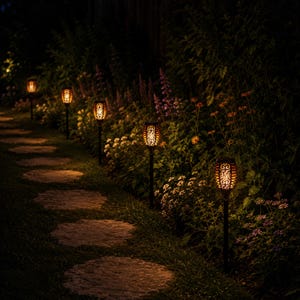 May include: A series of five black solar-powered garden stake lights with intricate cut-out patterns that emit a warm, flickering orange glow, positioned along a stone pathway lined with various green plants and small colourful flowers.