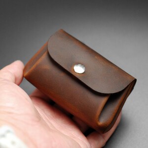 May include: A small, brown leather wallet with a rounded flap secured by a silver snap. The wallet has a textured appearance and is held in a hand against a dark gray background. The wallet appears to be empty.