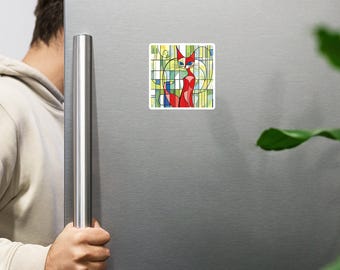 May include: A square magnet featuring a stylized red cat with blue eyes and a white face, set against a colorful geometric background of green, yellow, and blue lines. The magnet is attached to a silver refrigerator.