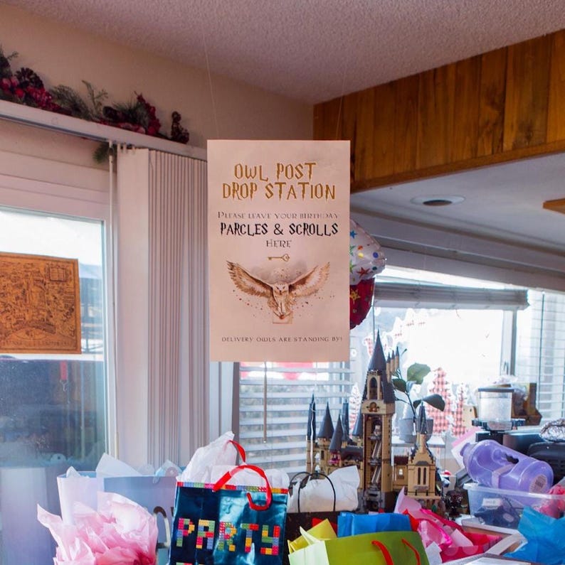 Puede incluir: Un letrero rectangular con el texto "OWL POST DROP STATION" y "PARCLES & SCROLLS HERE" cuelga del techo, con una ilustraci&oacute;n de un b&uacute;ho. Debajo, se ven bolsas de regalo coloridas y un castillo de juguete.