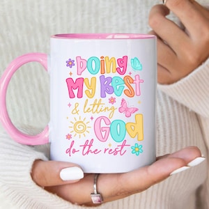 May include: White ceramic mug with a pink handle and interior. The mug features colourful text that reads "Doing My Best & Letting God Do The Rest" with butterfly and flower illustrations. A cheerful and inspirational design.