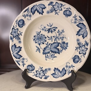 May include: Cream-colored ceramic plate with a blue floral design. The plate features a scalloped edge and a central floral arrangement. The design includes leaves, flowers, and fruit. The plate is displayed on a black stand.