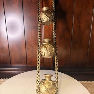 May include: A gold-colored, hanging decorative piece featuring three small, round vases. Each vase is encased in a woven basket-like design and suspended from a braided rope. The vases are displayed against a dark wood background.