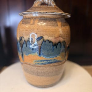 May include: A handcrafted ceramic jar with a lid. The jar is a warm tan color with blue and brown accents. The lid has a twisted rope-like handle. The jar is likely used for storage and decorative purposes.
