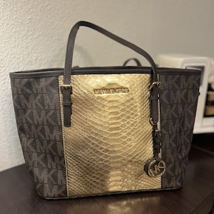 May include: A Michael Kors tote bag with a brown and gold color scheme. The bag features a gold snakeskin-like center panel, brown handles, and the brand's logo. The sides of the bag have a repeating MK pattern.