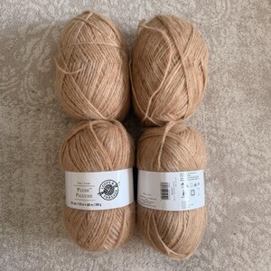 May include: Four tan yarn skeins arranged in a square. Two skeins are stacked on top of two others. The bottom two skeins have white labels with black text that reads "PLUSH PELUCHE". The yarn is a light brown color.