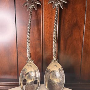 May include: A pair of silver-toned serving spoons. Each spoon features a palm tree design, with a detailed palm frond at the top and a textured trunk leading to the spoon bowl. One spoon has slots. The spoons are displayed on a white surface.