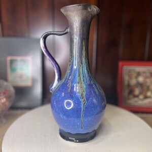 May include: A ceramic vase with a blue and black glaze. The vase has a bulbous body, a narrow neck, and a handle. The glaze has a dripped effect, with streaks of blue, black, and green. The vase is approximately 8 inches tall.