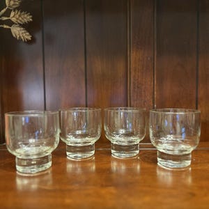 May include: Four clear, stemless shot glasses with a rounded bowl and a thick, flat base. The glasses are arranged in a row on a dark wooden surface. The background is a dark wooden cabinet.