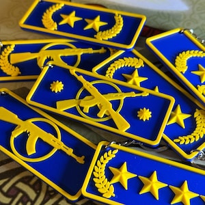 Puede incluir: Una colección de llaveros azules y amarillos. Cada llavero presenta un diseño rectangular con estrellas amarillas, coronas de laurel y rifles amarillos estilizados. Los llaveros están dispuestos, mostrando sus detalles.