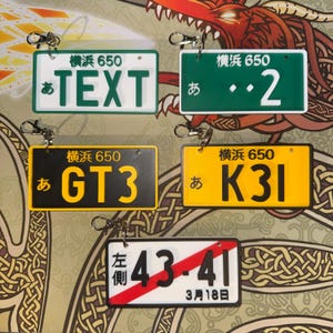 May include: A collection of miniature Japanese license plate keychains in various colors. The keychains feature text and numbers in Japanese, including "TEXT", "2", "GT3", and "K31". Each keychain has a silver clasp.