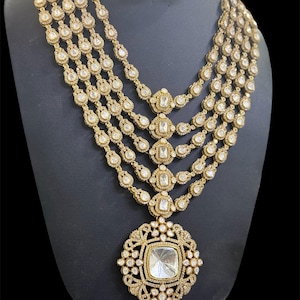 Peut inclure: Un ensemble collier et boucles d'oreilles de couleur dorée. Le collier présente plusieurs rangées de petites pierres transparentes et de plus grandes pièces centrales carrées. Le pendentif est un grand carré orné avec une pierre centrale. Les boucles d'oreilles reprennent le design.