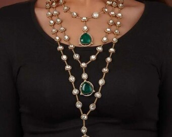 Op de afbeelding: Een ketting met drie strengen van goudkleurig metaal en heldere, ronde edelstenen. Elke streng heeft een grote, peervormige, groene edelsteen hanger. De ketting wordt gedragen over een zwarte top.