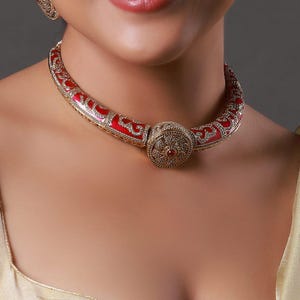 Puede incluir: Primer plano de una mujer que lleva un collar rojo y dorado y pendientes a juego. El collar tiene un diseño ancho y curvado con patrones intrincados y un colgante central circular. Los pendientes son circulares con detalles similares.
