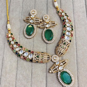 Puede incluir: Un conjunto de collar y pendientes dorados. El collar y los pendientes presentan grandes piedras ovaladas de color verde esmeralda rodeadas de piedras claras más pequeñas. El collar tiene un diseño curvo con una variedad de piedras de colores.