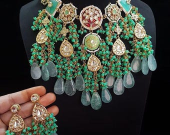May include: An ornate necklace and matching earrings set. The necklace features green and gold accents, with teardrop-shaped pendants and dangling green beads. The earrings mirror the necklace design, held in a hand against a black background.