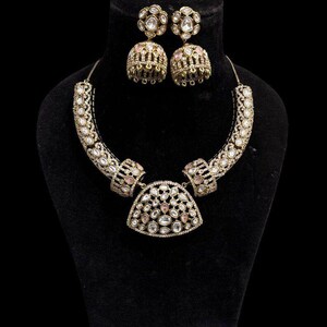 Puede incluir: Un conjunto de collar y pendientes dorados, exhibido sobre un busto negro. El collar presenta un diseño curvo con piedras preciosas claras y rosas. Los pendientes son redondos con detalles similares. La joyería es ornamentada y detallada.