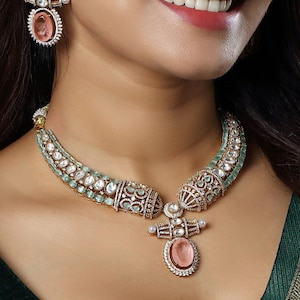 Puede incluir: Un conjunto de joyas dorado que incluye un collar y pendientes a juego. El collar tiene un colgante central con una gran gema rosa ovalada, rodeada de piedras transparentes más pequeñas. Los pendientes reflejan el diseño del colgante.
