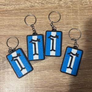 Peut inclure: Quatre porte-clés avec un motif rectangulaire bleu, une bande verticale blanche et une bordure noire. Chaque porte-clés est muni d'un anneau argenté. Le motif représente une lettre "I" stylisée.