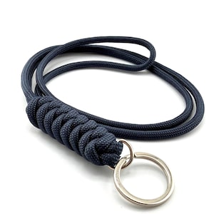 May include: A dark navy blue paracord lanyard featuring a tightly woven, braided section that transitions into a smooth cord loop, finished with a circular silver-toned metal split ring for attaching keys or other small items securely.