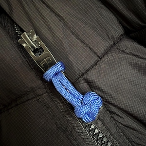 May include: A black quilted jacket featuring a metallic zip pull with the letters FC engraved on the surface. A bright blue braided cord is looped through the zip pull, ending in a decorative knot to assist with gripping the zip mechanism.