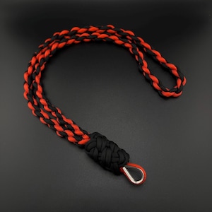 May include: A black and red braided lanyard with a metal clasp. The lanyard is made of a durable, woven material in a repeating pattern of black and red. The clasp is silver-coloured.