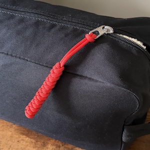May include: A black rectangular toiletry bag featuring a silver metal zip closure. A bright red paracord pull with a braided knot design is attached to the zip slider. The bag is constructed from a solid black fabric and includes a side handle.