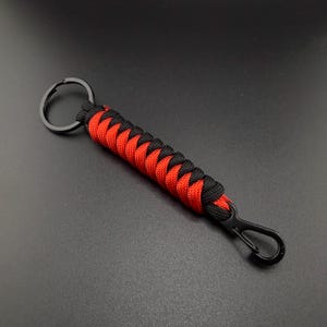 May include: A keyring featuring a braided paracord design in alternating black and red colours. The cord is attached to a circular black metal ring at one end and a black metal lobster clasp at the other end, creating a durable accessory.