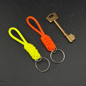 May include: Two paracord keychains, one in neon yellow and one in bright orange, each featuring a braided knot design and a circular metal split ring. A brass-coloured metal key with the word UNION embossed on the head is positioned nearby.