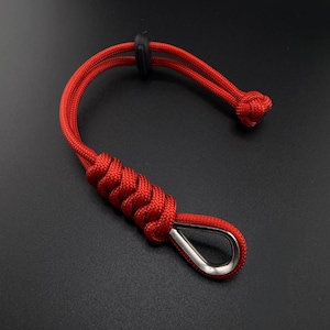 May include: A bright red paracord lanyard featuring a braided design, a metallic silver thimble at the loop end, and a black plastic sliding cord lock. The cord is arranged in a curved shape with a knotted end, showcasing a durable woven texture.