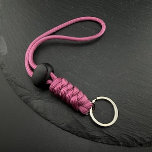 May include: A magenta paracord keyring featuring a braided lanyard loop, a black plastic cord lock, and a silver-toned metal split ring. The cord is woven into a tight, textured pattern leading to the circular metal attachment point.