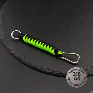 May include: A keyring featuring a braided paracord design in alternating black and neon green colours. The cord is attached to a circular metal ring on one end and a metal carabiner clip on the other. A circular label reads MARINE GRADE 316 A4 STAINLESS STEEL HARDWARE.