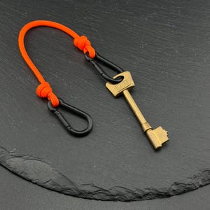 May include: A brass-coloured metal key with the word UNION embossed on the head, attached to a bright orange paracord loop. The cord features two black metal carabiners secured by knots, creating a functional keychain assembly for carrying keys.