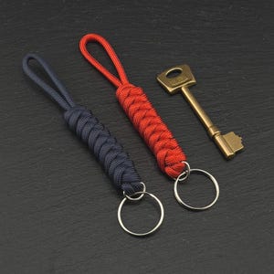 May include: Two paracord keychains, one in dark navy blue and one in bright red, each featuring a braided cord loop and a circular metal split ring. Beside them lies a single brass-coloured metal key with the word UNION embossed on the head.