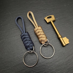 May include: Three items on a dark surface: two keychains with braided cord in navy and tan, and a brass key. The keychains have silver key rings. The brass key has the word "UNION" stamped on it.
