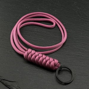 May include: A lanyard crafted from dark pink paracord featuring a braided section and a circular black metal key ring. The cord is looped to form a long strap, with the braided portion providing a textured grip near the attachment point.