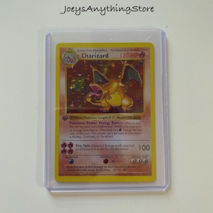 May include: A Charizard Pokémon trading card in a protective sleeve. The card features a fiery orange dragon with the name "Charizard" at the top. The card has a HP of 120 and includes text describing its abilities and attacks.