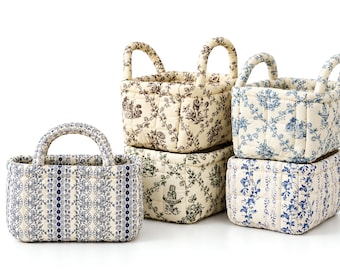 May include: Four rectangular fabric storage baskets with handles. One features a blue and white lace-like pattern. The others have quilted cream fabric with brown or blue toile-style prints depicting floral motifs and garden scenes.